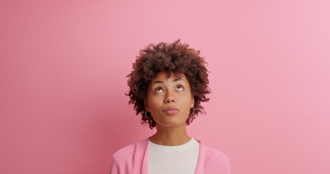 Satisfied dark skinned woman smiles gently looks on right left and upwards reads advertisement or promotional text makes surprised face poses against rosy background. Human emotions concept.
