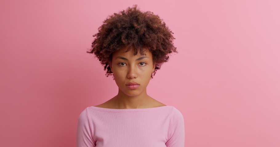 Slow motion beautiful young woman with Afro hair grimaces as if looking at something disgusting or ugly frowns face with aversion dislikes something unpleasant poses against pink background. Royalty-Free Stock Footage #1062011197
