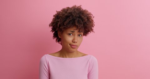 Slow motion beautiful young woman with Afro hair grimaces as if looking at something disgusting or ugly frowns face with aversion dislikes something unpleasant poses against pink background.