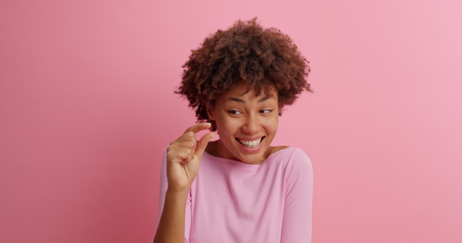 Young brunette young woman with curly Afro hair shows very tiny object shapes small item needs some more dressed in casual clothing poses against pink background. Minimum size. I need not much | Shutterstock HD Video #1062011203