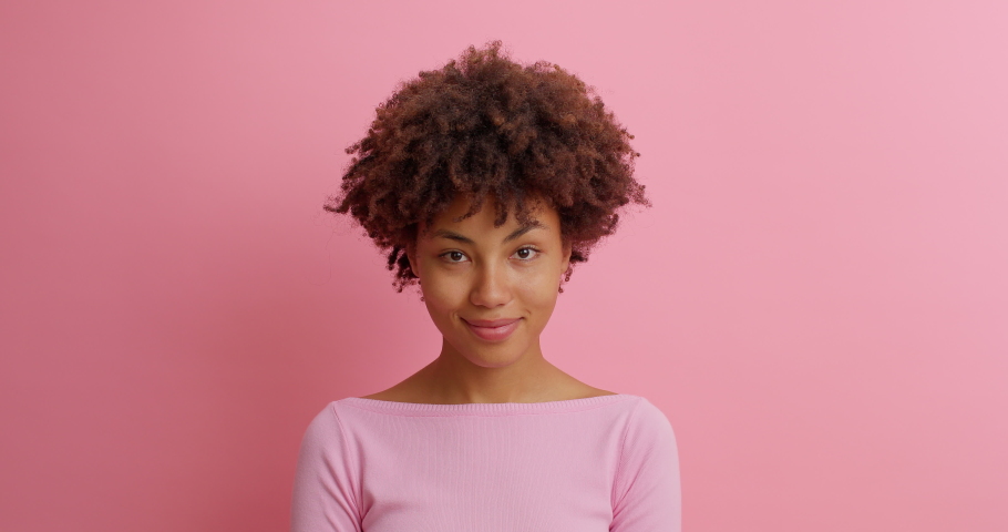 Happy pleased dark skinned woman looks on different sides with shy expression smiles broadly shows white teeth winks eye with flirt dressed in long sleeved jumper models over pink background | Shutterstock HD Video #1062011230