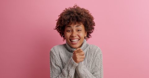 Happy dark skinned young woman makes triumph gesture says yes I did it makes victory dance glad to achieve great results dressed in knitted sweater poses against rosy background. Success concept