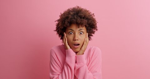 Impressed embarrassed dark skinned woman stares at camera with bugged eyes keeps jaw dropped sees something unbelievable holds breath from amazement isolated on pink background. Reaction concept