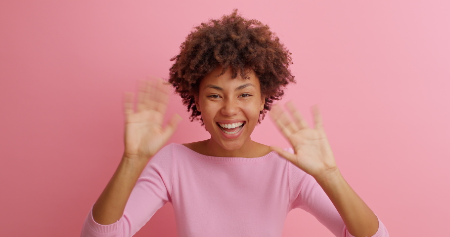 Happy friendly young woman waves palm in hello gesture welcomes someone with hospitable expression expresses positive emotions dressed casually isolated over pink background. Hi to all of you