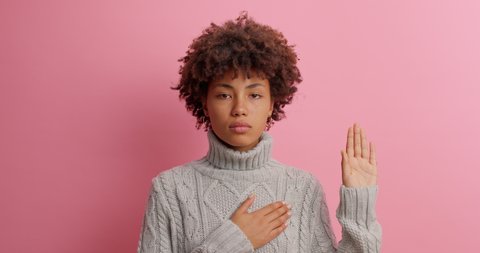 I swear to be honest! Serious dark skinned young woman makes promise gesture assures you to tell only truth dressed in knitted sweater isolated over pink background. Oath loyalty pledge concept