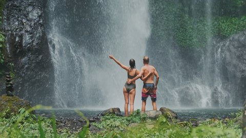 Rear view of young traveling couple standing in front of waterfall with their hands raised. Woman and man tourists with their arms outstretched looking at waterfall.