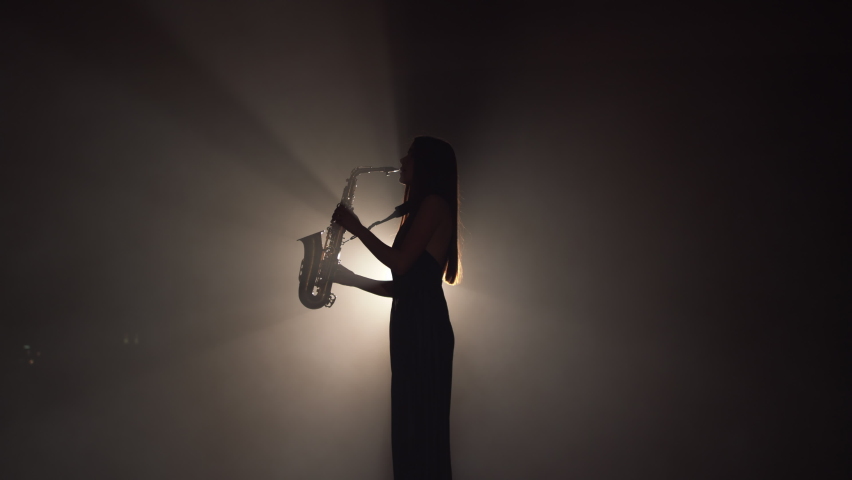 Young beautiful girl in a dark dress plays on a golden shiny saxophone on stage. Dark studio with smoke and stage lighting. Hands and saxophone close up. Side view.Slow motion video