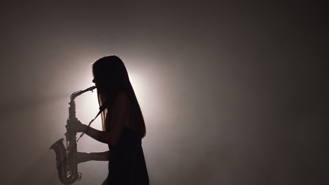 Young beautiful girl in a dark dress plays on a golden shiny saxophone on stage. Dark studio with smoke and stage lighting. Hands and saxophone close up. Side view.Slow motion video