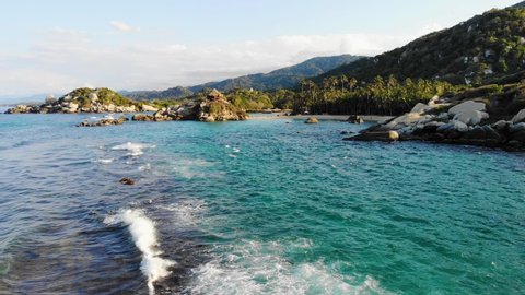 Aerial view low over waves and shallow, blue sea, on a rocky coastline, in Tayrona national natural park, during golden hour, in the Caribbean region of Colombia - dolly, drone shot