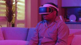 Close up mixed race young man wearing virtual reality headset sitting at home neon light. Fun game. VR glasses. Social network robot. Slow motion