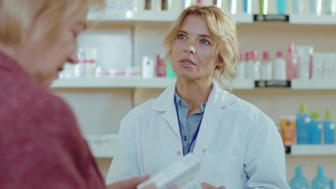 Attractive blond woman pharmacist serving a customer in a drugstore. Conversation pharmaceutical client. Seller commercial health care buyer uniform. Slow motion