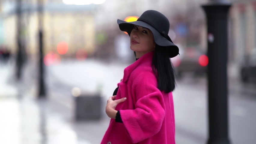 Portrait of a pretty black-haired woman in a pink coat and black hat walking on a blurry background of a city street near the road. Smiling and posing. | Shutterstock HD Video #1062023335