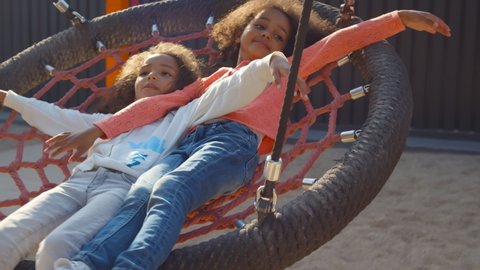Smiling little african girls riding on swing with net outdoors. Portrait of adorable afro-american toddler sister lying on net swing and playing in park playgroundの動画素材