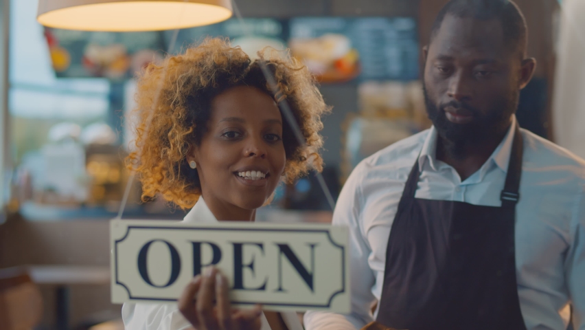 African cheerful small business owners smiling and turning open sign on cafe glass door. Happy successful afro-american couple of entrepreneurs opening new bistro restaurant