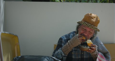 Homeless senior man with dirty clothes and old blanket wearing medical protection mask getting food from kind people and he is eating bread with happiness at the sidewalk