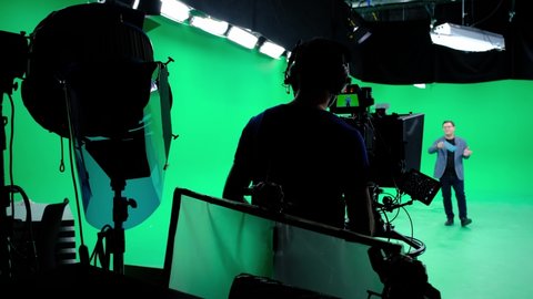 The cameraman follows the announcer in a professional green screen studio. The professional looks all the shooting equipment; lights, camera, green screen, monitors, filters
