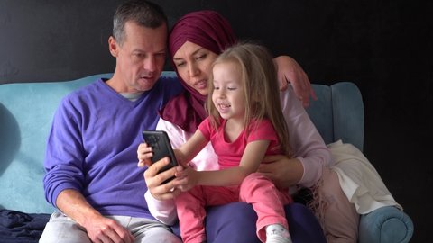 Happy Traditional Muslim family-father, mother in hijab and baby girl sitting together at home on the sofa. Coronavirus, Quarantine, Social Distancing, Self-Isolation, Lockdown