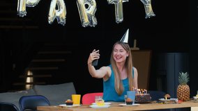 Self isolation birthday. Happy Caucasian young woman talks to friends, blows on cake on smartphone video call at home.