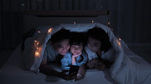 Asian family Father, mother and daughter spending time with mobile devices on the bed under the blanket at home night shots. Connection, Communication, Entertainment, Fun from smartphone