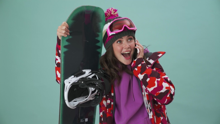 A pleased young woman snowboarder is talking on her smartphone standing isolated over a blue background in the studio