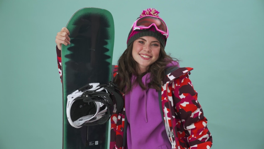 A happy young woman snowboarder is doing thumb-up gesture standing isolated over a blue background in the studio