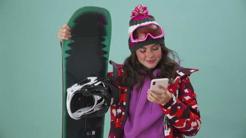 A positive young woman snowboarder is using her smartphone standing isolated over a blue background in the studio