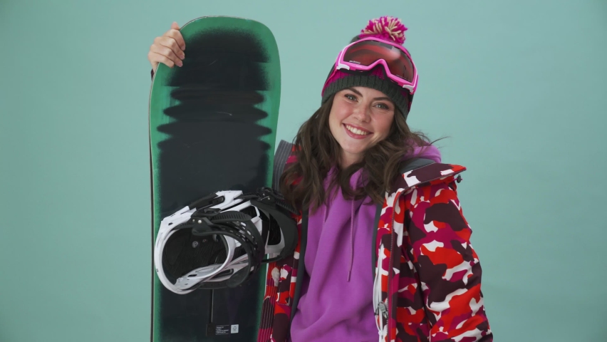 A smiling young woman snowboarder is standing isolated over a blue background in the studio