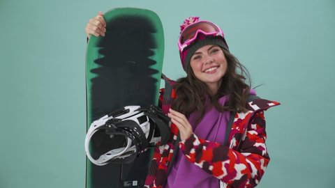A smiling young woman snowboarder is standing isolated over a blue background in the studio