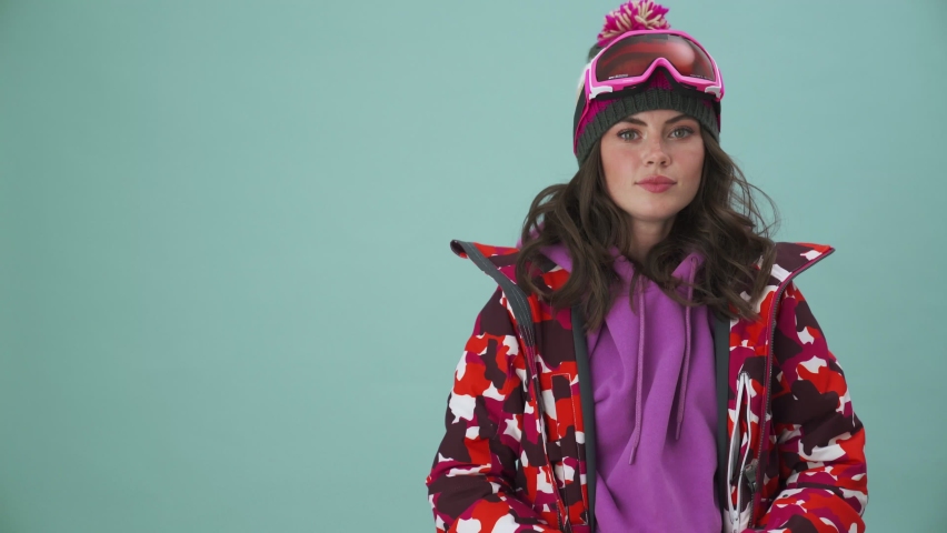 A beautiful young woman snowboarder is holding a cup of coffee standing isolated over a blue background in the studio