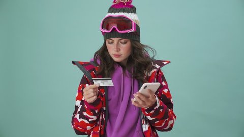 A beautiful young woman snowboarder is using her smartphone and credit card standing isolated over a blue background in the studio