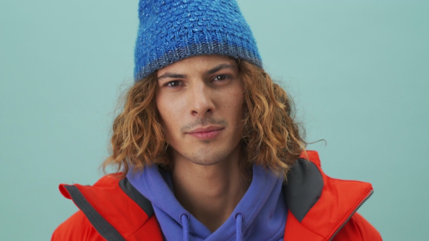 A handsome snowboarder is winking while looking to the camera standing isolated over a blue background in the studio