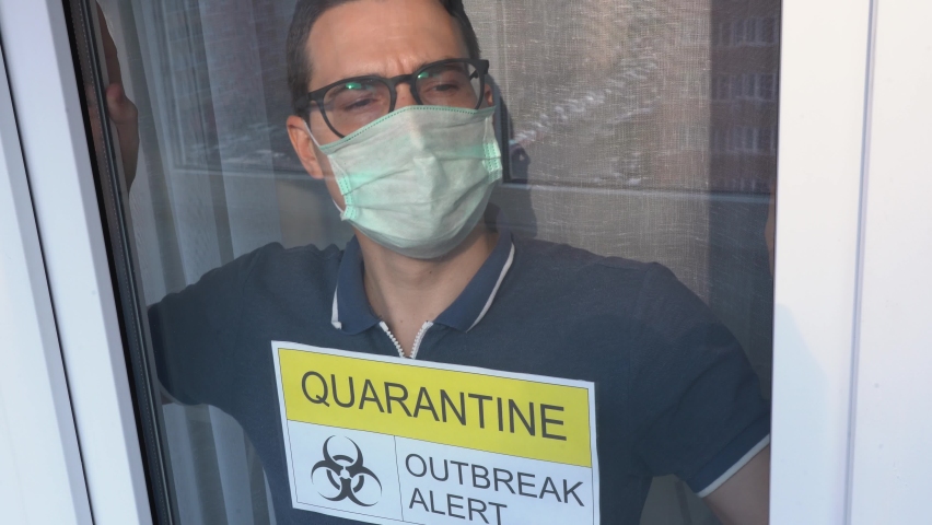 Worried Isolated infected sick young man wearing blue T-shirt, protective turquoise face mask, leans glass, looks out from hospital ward close up. Yellow white warning sign inscription glued on window | Shutterstock HD Video #1062050086