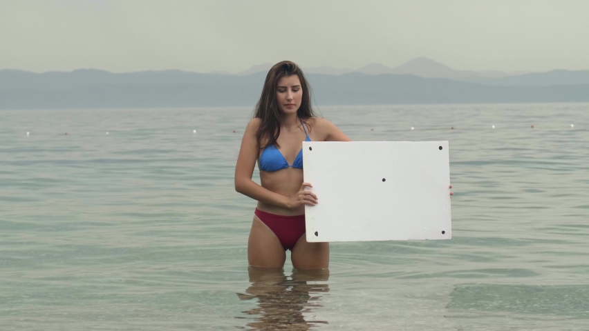 Young woman in bikini holding a white billboard in the sea against a blue sky background, is surprised what she sees on the billboard. Creative people can place any desired image on the whiteboard.