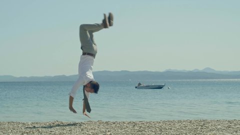 A young man in a suit, wearing a tie and doing various jumps while running on the sea beach at sunset. He does somersaults and poses for the camera.