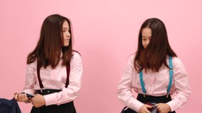 Beautiful little twin schoolgirls having fun fighting with their backpacks. Over pink background studio video.