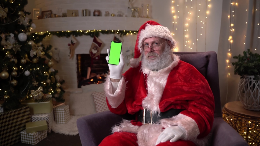 Happy Santa Claus sitting in chair near christmas tree and fireplace, showing mobile phone with green screen. Christmas spirit, holidays and celebrations concept 4k footage Royalty-Free Stock Footage #1062056623