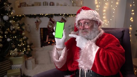 Happy Santa Claus sitting in chair near christmas tree and fireplace, showing mobile phone with green screen. Christmas spirit, holidays and celebrations concept 4k footage
