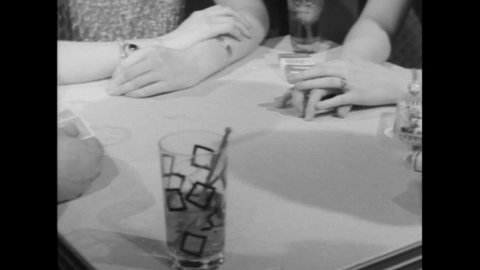 CIRCA 1960s - Two couples play strip poker and a blonde woman loses, takes off her top, sips her drink and lights a cigarette, in her black bra.