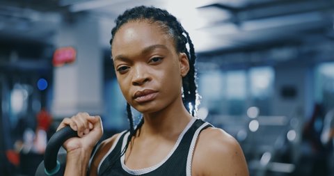 Portrait of powerful african american strong sportswoman with braids holding dumbbells looking confident at camera posing inside gym. Sportswoman. Attractive look. Active lifestyle.
