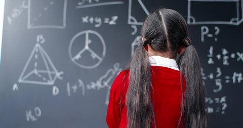 Rear of Caucasian small teen girl writing math equations on blackboard at class, turning and smiling to camera. Back view on little schoolgirl at chalkboard doing mathematics. School concept.