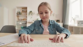 Lockdown of blond-haired Caucasian girl is sitting at kitchen table in front of computer, typing on keyboard and showing home task to the teacher by video link