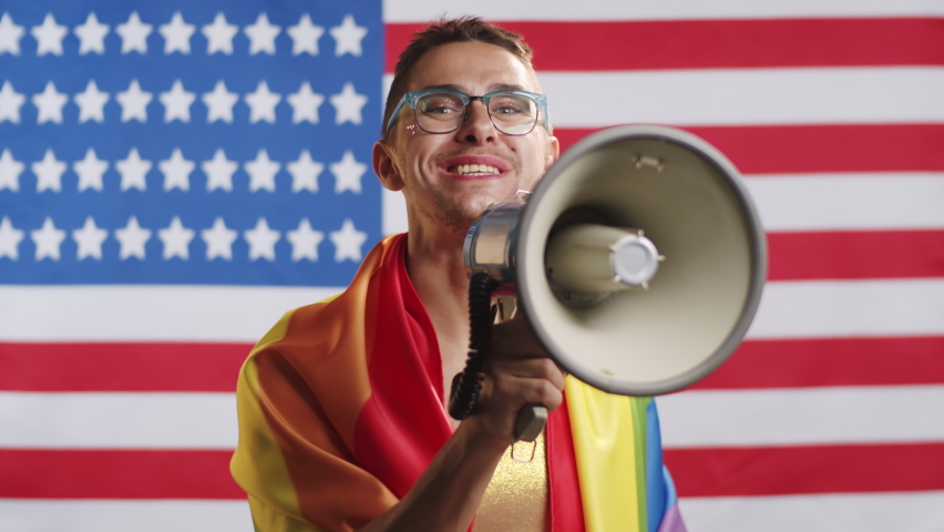 Excited gay man looking at camera, raising arm and yelling into loudspeaker while giving public speech against American flag during press conference