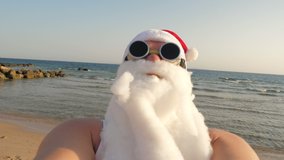 Santa Claus blogger. Santa Claus takes selfie. Funny Santa, in sunglasses, takes selfie video on beach by the sea, against palm trees backdrop. Santa summer vacation.
