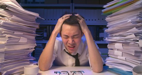 Stressed asian business man is sitting at desk covered with stack of paperwork and having nervous breakdown while working hard at late night. Angry alone male is frustrating and screaming in anger.