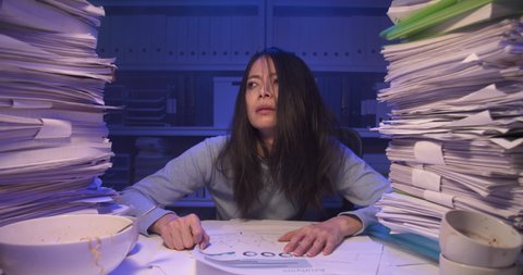 Sleepy exhausted asian business woman looking at document and mess of her hair while working hard at late night. Stressed alone girl throw papers and lay down on desk covered with stack of paperwork.