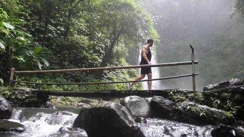 A young, fit, white man is crossing a handmade bamboo bridge that goes over a river in Bali. Beautiful and big waterfall in the background. Slow motion.