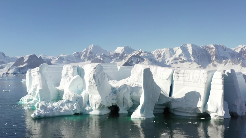 4K Aerial Landscape of snowy mountains and icy shores in Antarctica. Beautiful blue iceberg with mirror reflection floats in open ocean. Glaciers of a harsh continent. Royalty-Free Stock Footage #1062100405