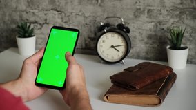 a man at a white Desk is sitting with a mobile phone in his hands with a green screen. swipes and taps on the smartphone's chromakey screen in close-up. ready-made video for inserting and keying