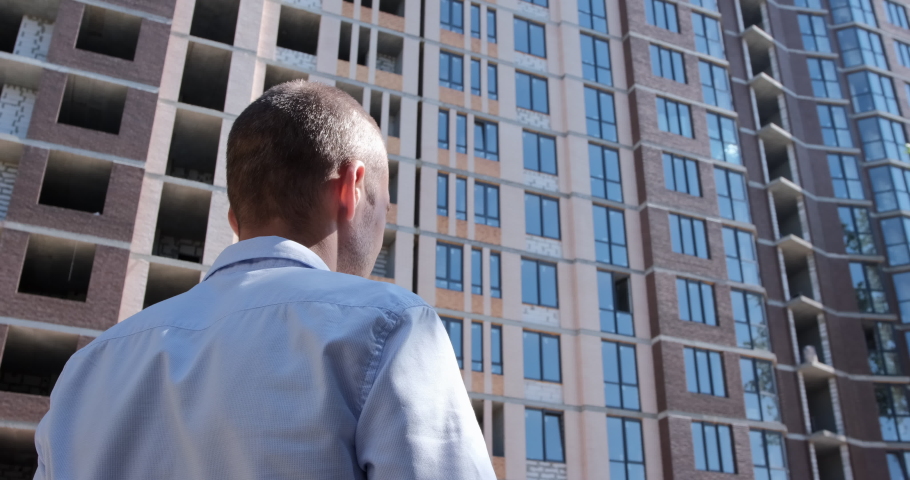 A man in a shirt puts a protective white helmet on his head and looks at a residential building under construction. Safety engineering at construction sites. The camera shoots a person from the back. Royalty-Free Stock Footage #1062104281