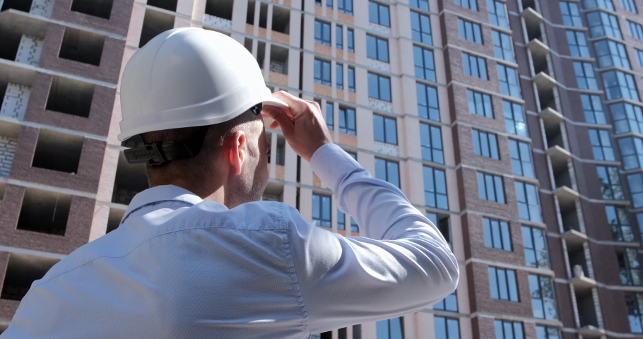 A man in a shirt puts a protective white helmet on his head and looks at a residential building under construction. Safety engineering at construction sites. The camera shoots a person from the back. | Shutterstock HD Video #1062104281